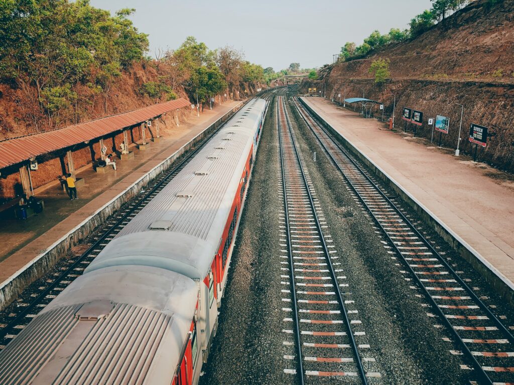 Konkan Railway track winding through lush green Western Ghats during monsoon. High-angle view of an Indian train station with a red tile roof in the Sahyadri mountains. Scenic railway route in India with dense forest and laterite soil cuttings. Lush landscape along the Konkan Railway in Maharashtra or Goa. Small red-roofed railway station surrounded by tropical green foliage. Monsoon travel photography: Indian train journey through deep green hills. Railway engineering marvel through the challenging, forested terrain of the Western Ghats. Indian Railway travel photograph, high-angle view of a secluded mountain station. Panoramic view of the Konkan coastal railway line and valley. Train track on a hill with vibrant green foliage, India.