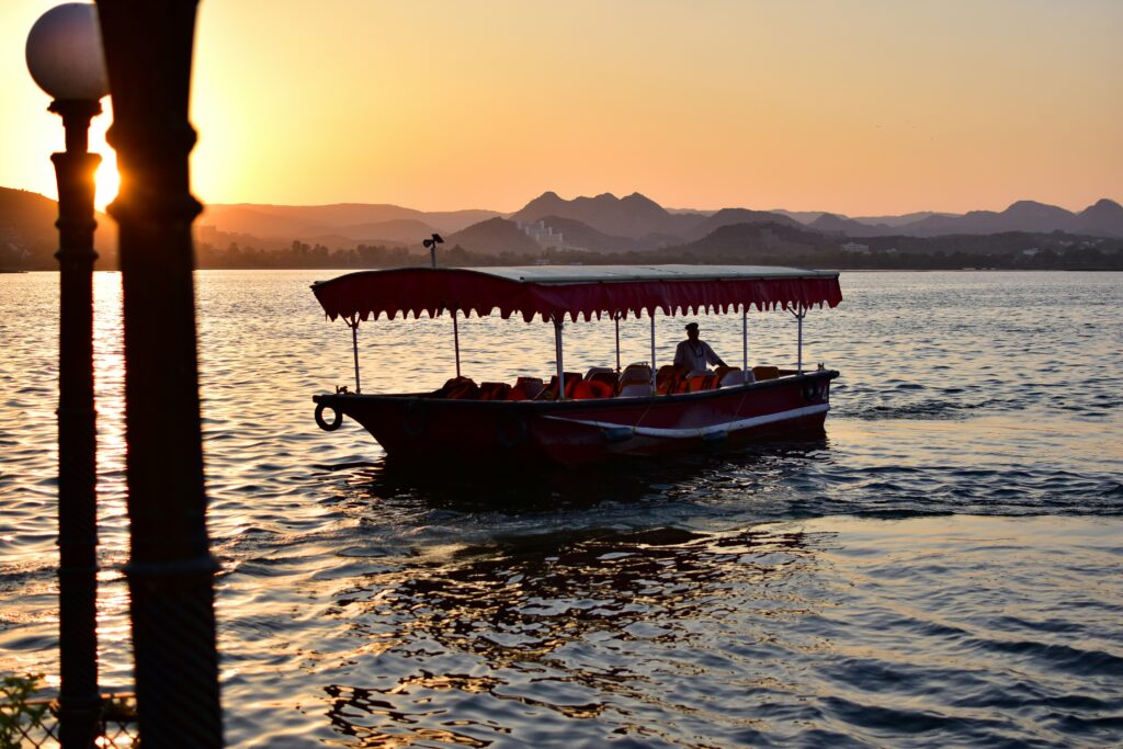 sunset boat ride lake pichola udaipur rajasthan india lake tourism photography udaipur city of lakes scenic view silhouette of boat on water at dusk golden hour lake pichola rajasthan tourist boat water reflections mountains udaipur travel destination boat safari peaceful evening scene indian lake udayaditya barua lake photography boatman rowing serene water orange sky